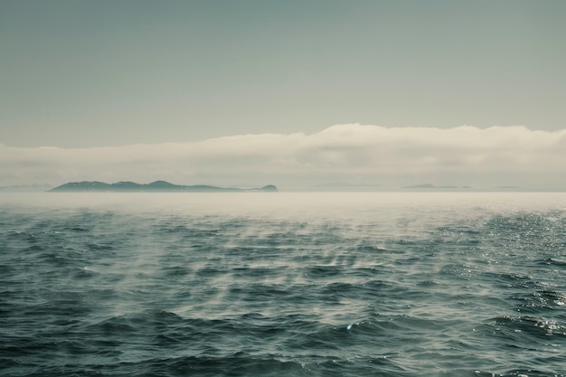 A vast body of water with a small island barely visible in the distance on a misty morning A misty morning on the open ocean with fog enveloping distant islands on the horizon