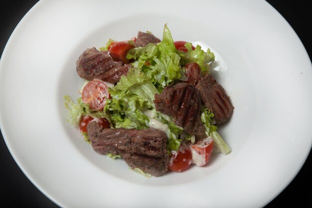 Veal salad, salad with large slices of veal and fresh vegetables on a plate on a black background.