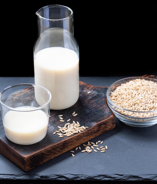 Vegan dairy free oat milk in glass bottle and glass and oat seeds on glass bowl on dark background