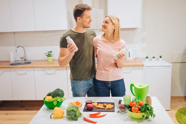 Vegan happy loving couple holds bottles with natural smoothie while cooking raw vegetables in white kitchen. Diet detox. Happy family. Raw food diet. Vegetarian food