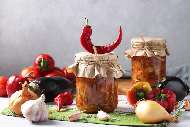 Vegetable salad with eggplant, carrot, peppers, garlic and tomatoes in glass jars on gray background. Autumn still life