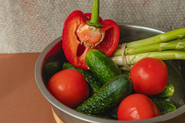 Vegetables lie in a metal bowl tomatoes asparagus cucumbers red bell peppers on a wooden board and brown background back gray background place for text