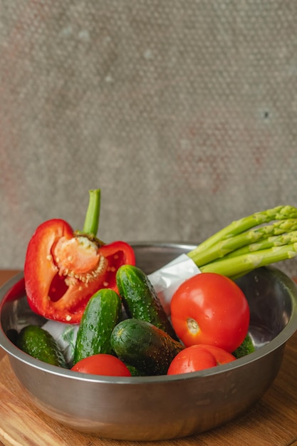 Vegetables lie in a metal bowl tomatoes asparagus cucumbers red bell peppers on a wooden board and brown background back gray background place for text