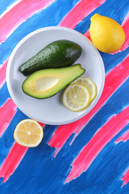 Vegetarian food concept Two halves of a green fresh ripe avocado and a juicy yellow lemon on a white plate on a bluepink background Closeup