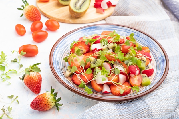 Vegetarian fruits and vegetables salad of strawberry, kiwi, tomatoes, microgreen sprouts on white concrete surface and linen textile