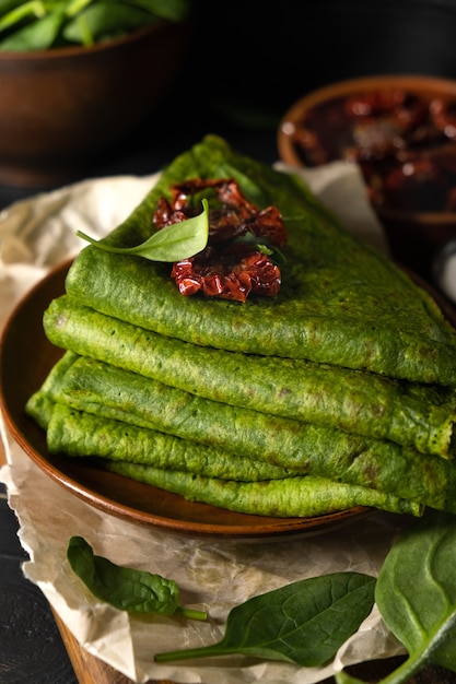 Vegetarian green pancakes decorated with dried tomatoes. Spinach pancakes in a brown plate on dark wooden background.