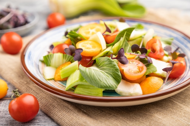 Vegetarian salad of pac choi cabbage, kiwi, tomatoes, kumquat, microgreen sprouts on gray wooden surface. Side view, selective focus.