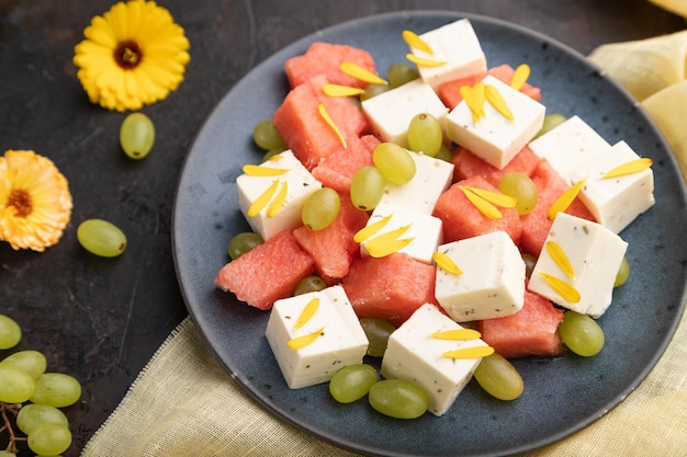 Vegetarian salad with watermelon, feta cheese, and grapes on blue ceramic plate on black concrete background and yellow linen textile. Side view, close up, selective focus.