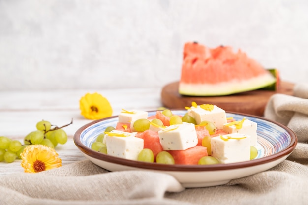 Vegetarian salad with watermelon, feta cheese, and grapes on blue ceramic plate on white wooden background and linen textile. Side view, close up, selective focus.