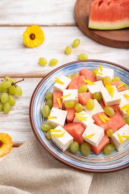 Vegetarian salad with watermelon, feta cheese, and grapes on blue ceramic plate on white wooden background and linen textile. Side view, close up, selective focus.