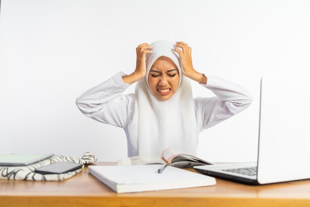 Veiled high school student at desk in stress pose using laptop on isolated background