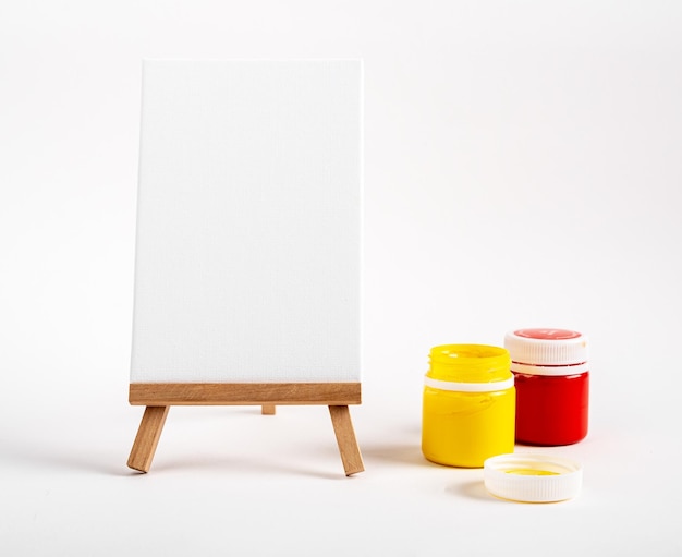 Vertical canvas blank paper on wood easel gouache paints in jars red and yellow