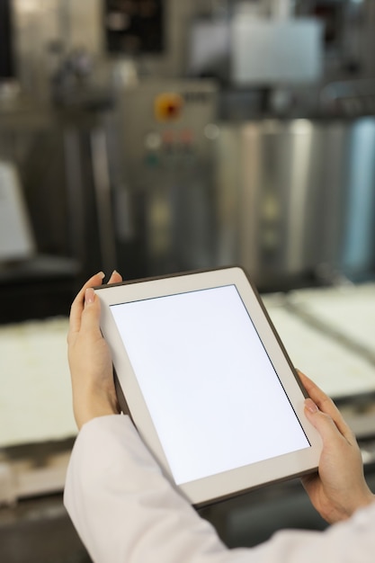 Vertical close up of female hands holding digital tablet with blank white screen against factory equipment in background, copy space