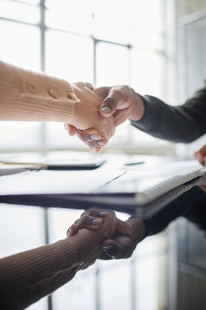 Vertical closeup of two people shaking hands over meeting table in office with mirror reflection