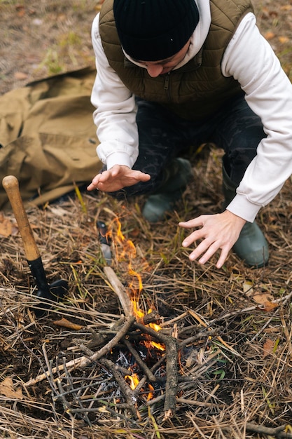 Vertical cropped shot of frozen traveler man warming hands over fire at outdoors on overcast cold day