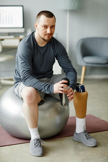 Vertical full length portrait of man with prosthetic leg sitting on fitness ball and holding water b