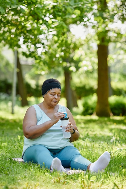 Vertical full length portrait of overweight black woman working out outdoors and drinking water to k