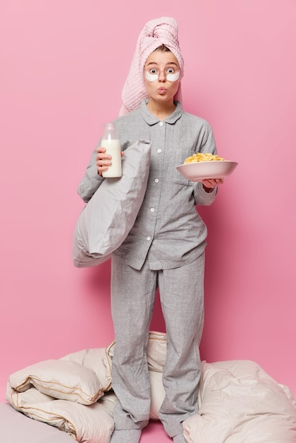 Vertical full length shot of young pretty woman keeps lips folded dressed in grey pajama holds pillow bottle of milk and cereals awakes in morning and has breakfast isolated over pink background.