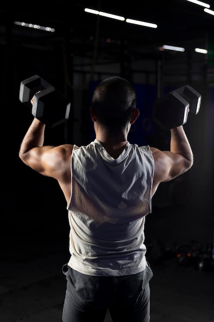 Vertical image of a muscular man from the back pressing with a pair of dumbbells