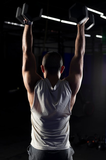 Vertical image of a muscular man from the back pressing with a pair of dumbbells