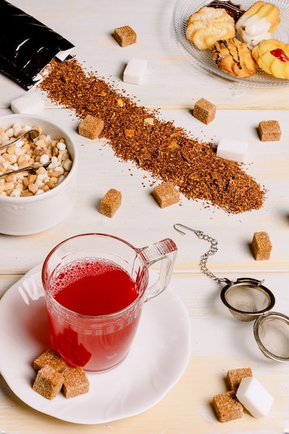 Vertical image of a red tea cup on weathered white wooden planks with sugar cubes scattered on the table and plate with tea pastries in the upper corner