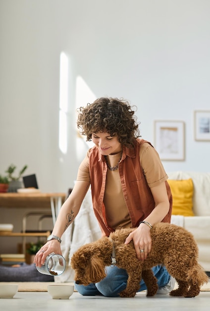 Vertical image of young woman feeding her little dog pouring food in bowl in the room