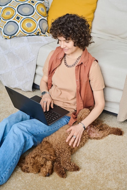 Vertical image of young woman resting at home with dog and using laptop