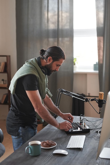 Vertical long shot of young bearded man adjusting audio mixer setting sound in home studio before st