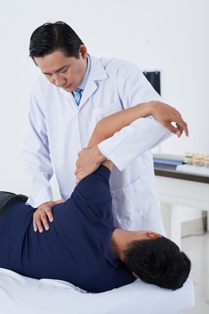 Vertical medium long shot of professional physical therapist working with unrecognizable young male patient recovering his functional abilities after severe injury