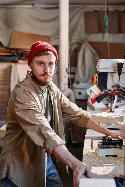 Vertical medium portrait of young caucasian male carpenter wearing red knit cap working with drill p