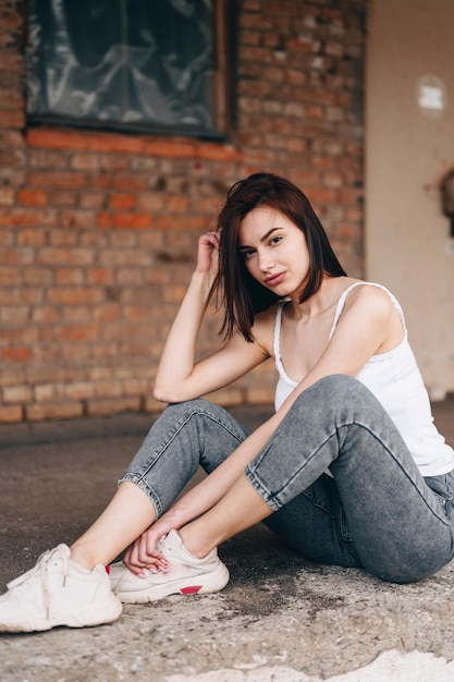 Vertical photo of a brunette woman in jeans sits on the ground The girl straightens her hair