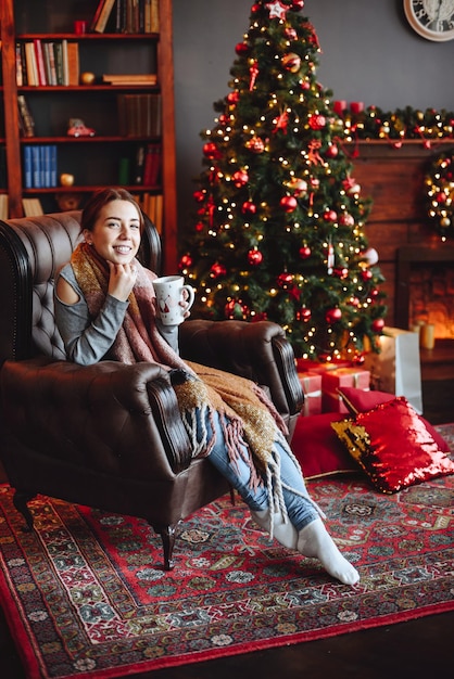 Vertical Photo of Cute Smiling Woman Sitting in an Armchair with Cup in Her Hands Looking at Camera