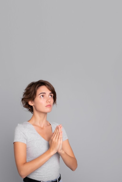 Photo vertical photo of cute young woman stands in meditative pose enjoys peaceful atmosphere hands in praying gesture and asking help isolated over gray background peace in soul