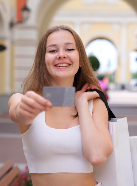 Vertical photo of Happy blurred young woman is showing holding credit bank card in hand and smiling during shopping with shopping bags in mall