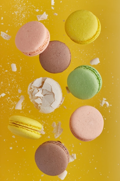 Vertical photo of macaroon. Colorful cake macaron with pastel tones and white cracked macaroon with crumbs in chaotic levitation on an yellow wall. Top view of almond cookies.