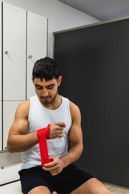 Vertical photo man young adult mixed race in the gym locker room sitting putting red boxing bandages on his hands Copy space Sports and recreation concept