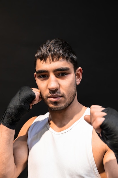 Vertical photo portrait of man young adult mixed race boxer looking at camera with fists with black boxing bandages raised on black background Sports recreation concept