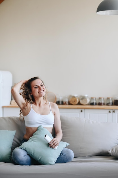 Vertical photo of young woman sitting on sofa and satisfactorily fixing her hair with her hand