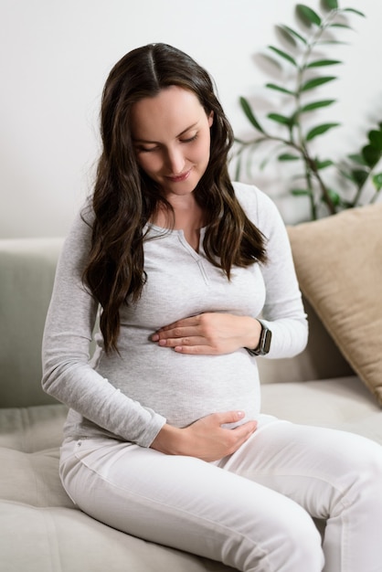 Vertical portrait beautiful pregnant caucasian woman sitting on couch touching pregnant belly, happy expecting concept, maternity