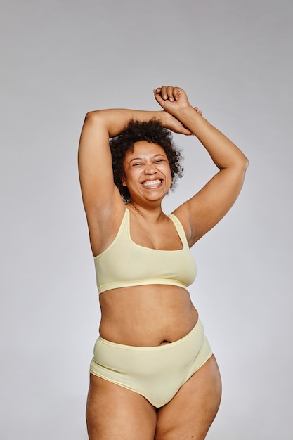 Vertical portrait of carefree black woman wearing underwear and laughing happily against grey backgr