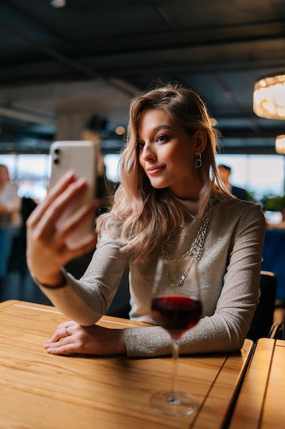 Vertical portrait of charming blonde female sitting at table with glass of red wine using cellphone live on social media writing stories taking selfies