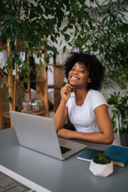 Vertical portrait of cheerful AfricanAmerican young woman holding pen in hand smiling looking at camera sitting at desk in light home office room