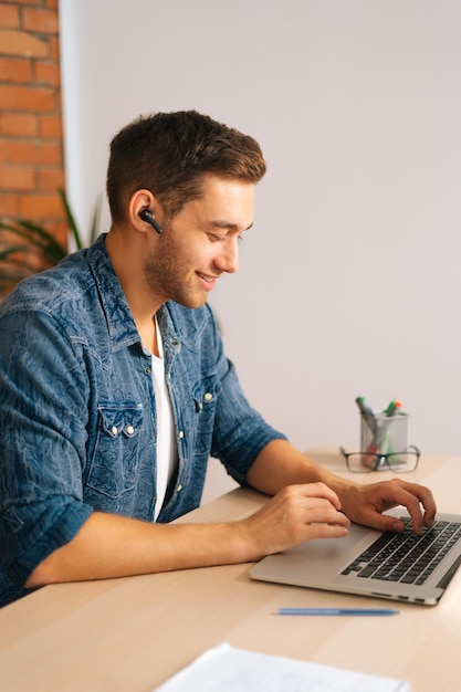 Vertical portrait of cheerful handsome freelance programmer male in wireless earphone working on laptop computer sitting at desk at home office