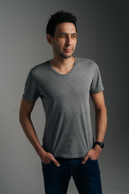 Vertical portrait of confident young man in gray T-shirt standing with with hands in pockets on dark isolated background. Studio shot of Caucasian male wearing casual clothes posing looking away