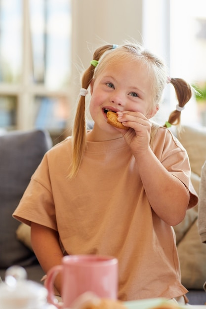 Vertical portrait of cute girl with down syndrome eating cookie while enjoying tea with family at home