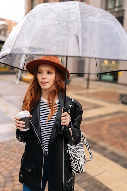 Vertical portrait of elegant redhead young woman in stylish hat holding in hand cup with hot coffee standing with transparent umbrella in cold autumn rain