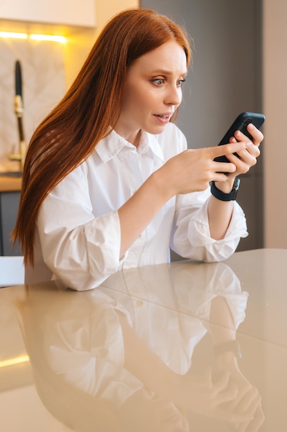 Vertical portrait of happy attractive redhead young woman checking mobile phone content sitting at