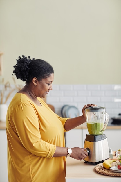 Vertical portrait of overweight black woman using blender while making healthy smoothie at home kitc