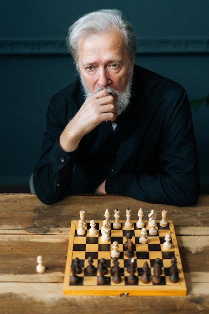 Vertical portrait of sad thoughtful bearded mature male thinking about chess move sitting at home and playing chess alone