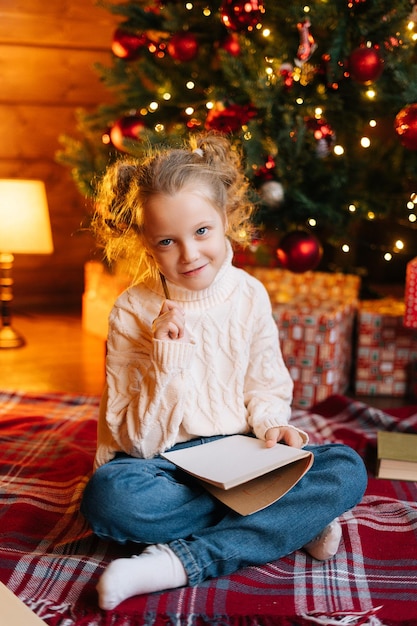 Vertical portrait of smiling little blonde curly girl writing letter to santa claus sitting on floor